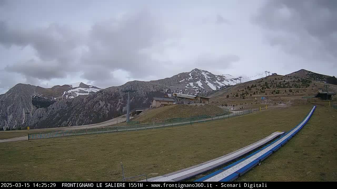 A grassy ski resort area with snow-capped mountains in the background under an overcast sky.
