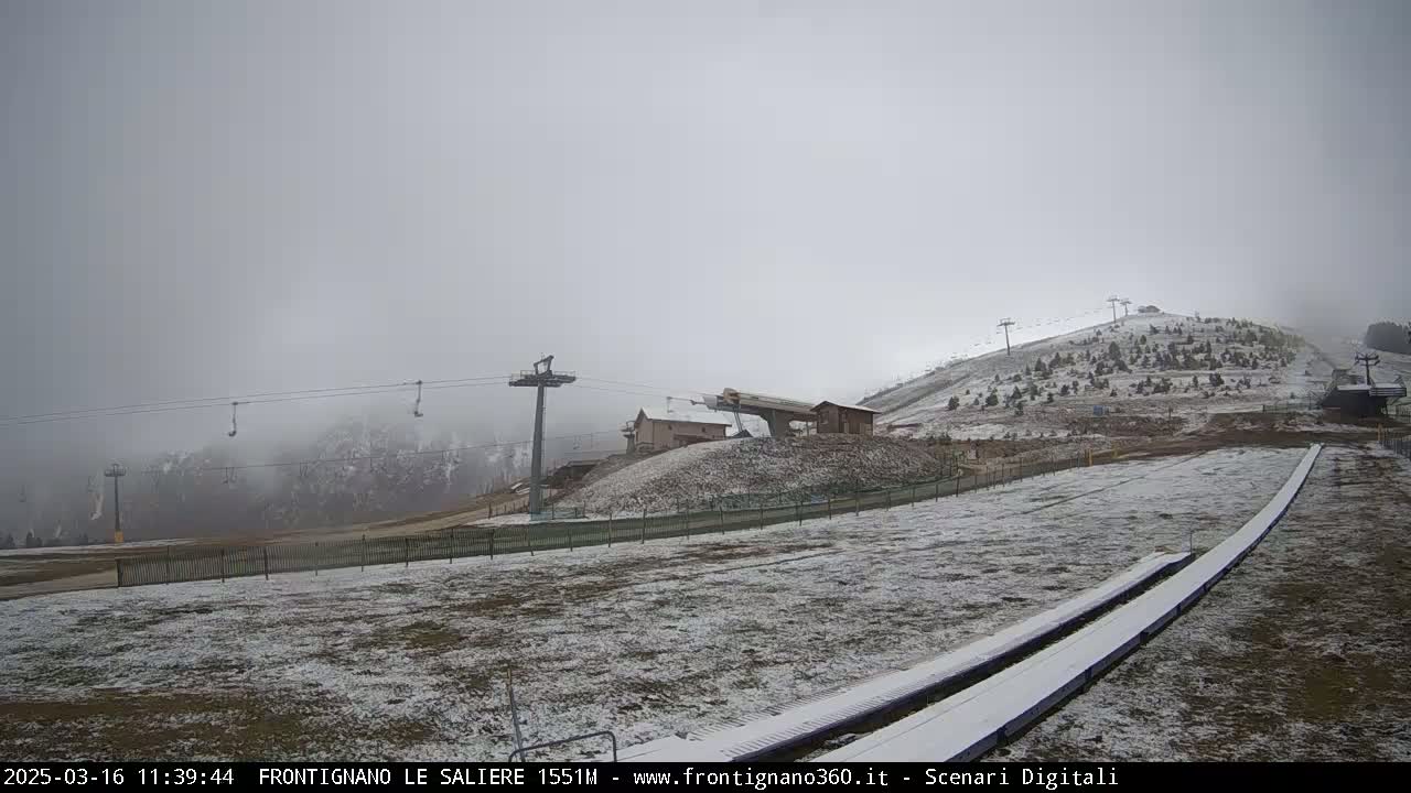 A lightly snow-covered ski slope with chairlifts and a ski lift station is visible under a foggy sky.