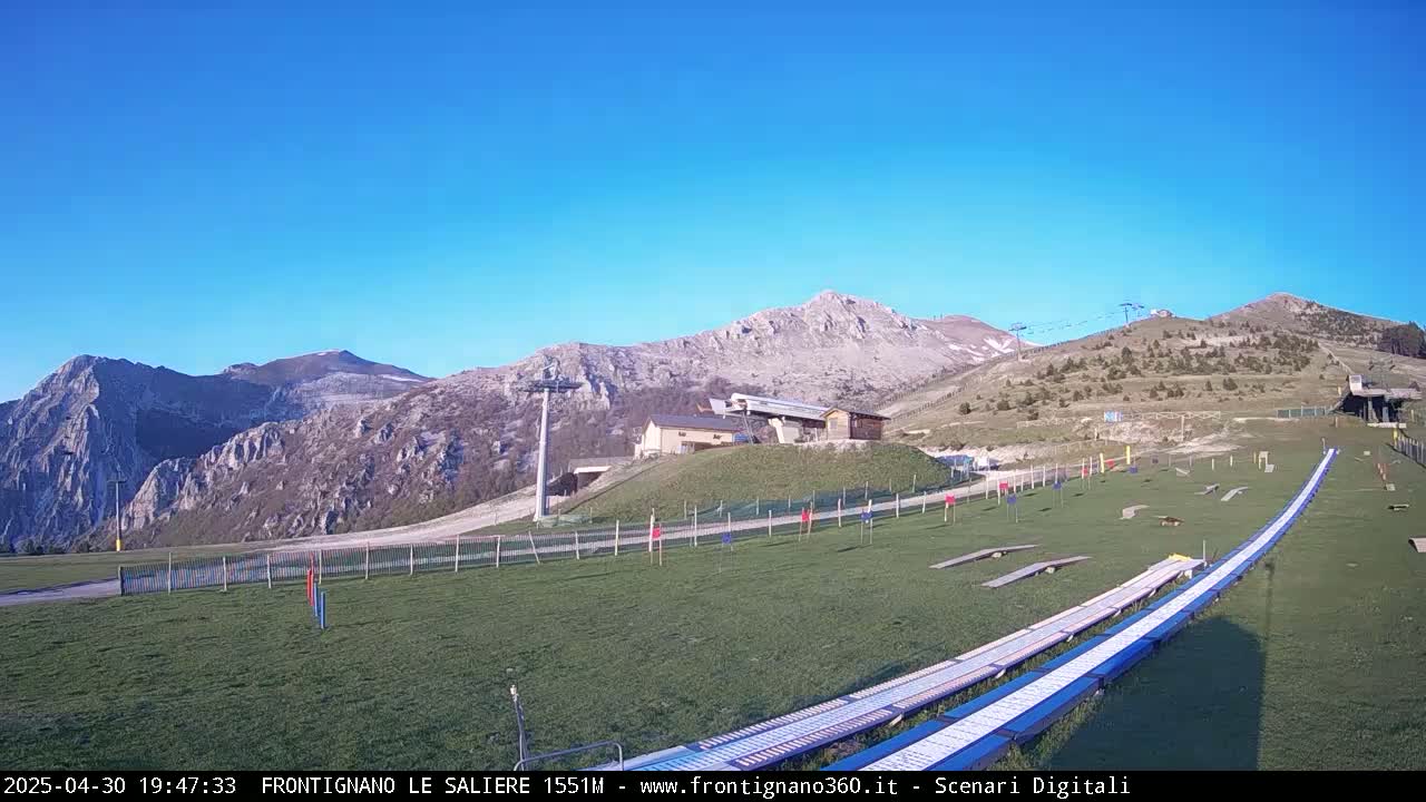 A grassy ski slope with a ski lift and a mountain range in the background under a clear blue sky.