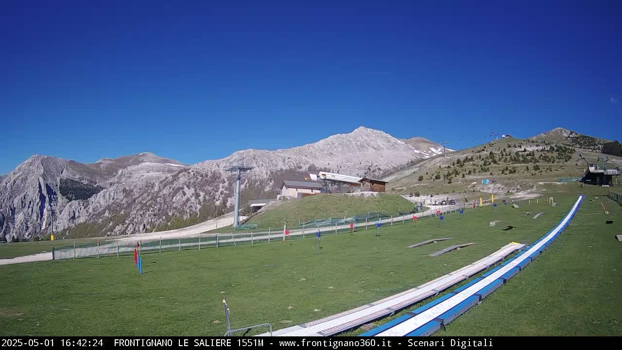 A grassy area with a long, white slide stretches towards a mountain range under a clear blue sky.