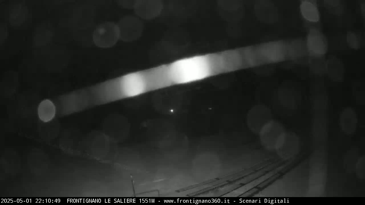 A blurry, nighttime, outdoor scene shows a bright streak of light across a dark area with rain or snow falling.