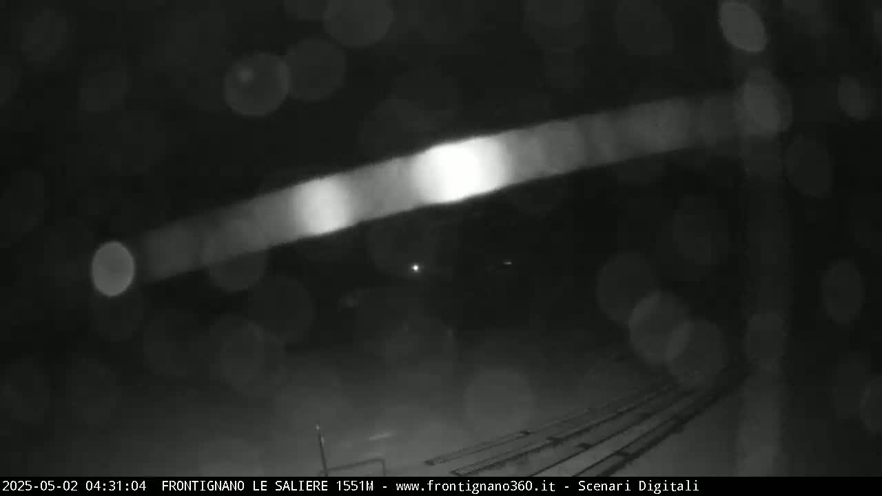 A blurry nighttime image shows a brightly lit horizontal streak in the darkness, possibly from headlights, with rain or snow visible as blurry spots.