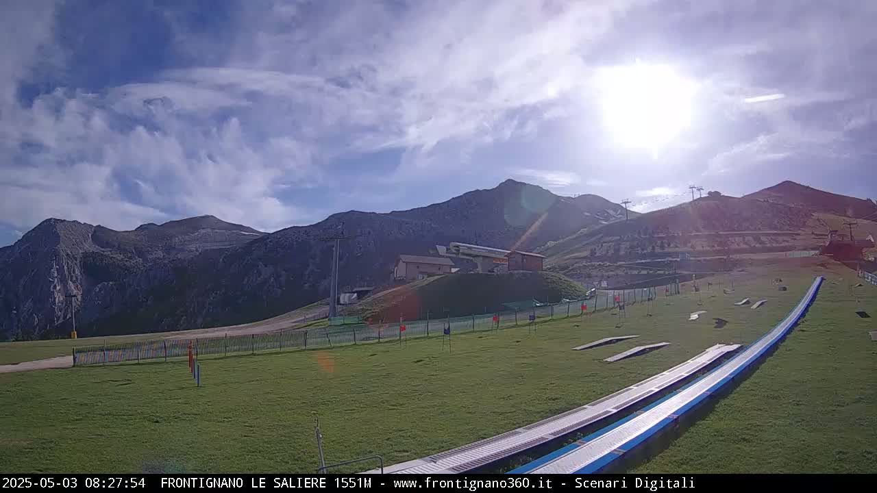 A sunny day reveals a grassy mountainside with ski lifts, a small building, and two long, blue ramps extending into the foreground, under a partly cloudy sky.