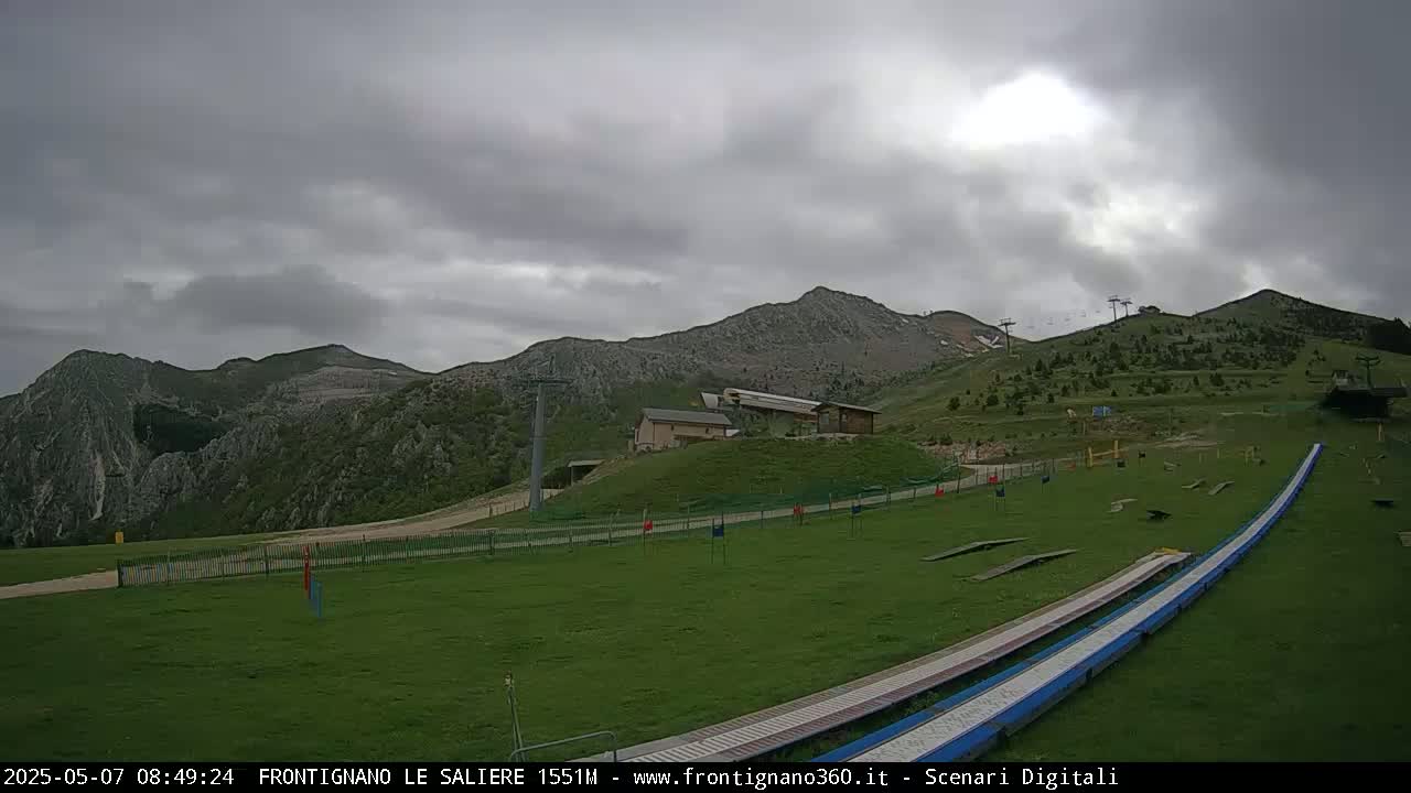A grassy hillside with a ski lift and two long, flat ramps leading down, set against a backdrop of mountains under an overcast sky.