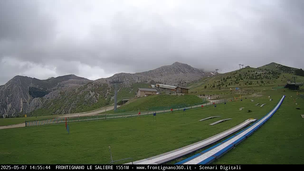 A grassy mountain slope with ski lifts, a small building, and two long, low ramps under an overcast sky.