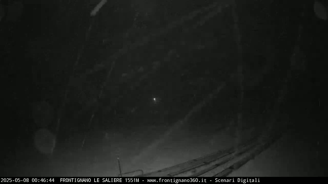 A nighttime, snowy scene shows a dark landscape with indistinct objects and a faint light in the distance.