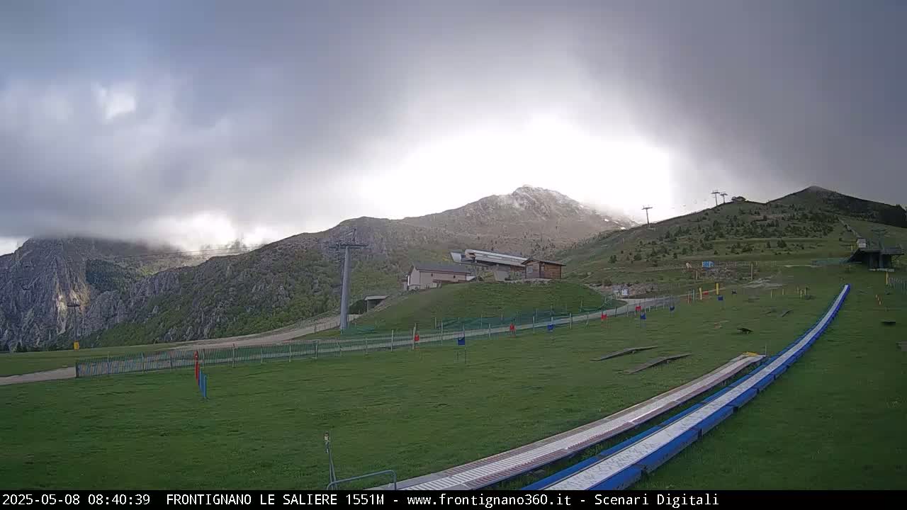 A grassy mountainside with a ski lift and a long, blue track extends toward a partly cloudy sky.