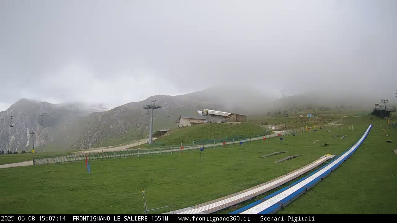 A foggy mountain landscape shows a grassy area with ski lifts, a small building, and a long, curved track.