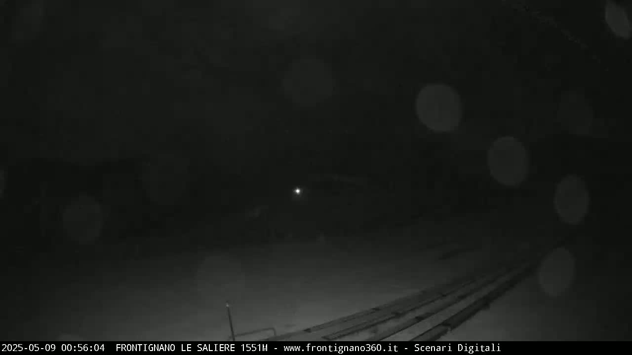 A nighttime scene shows snow falling on a dark, mostly empty area with a few faint lights and long, parallel lines on the ground.