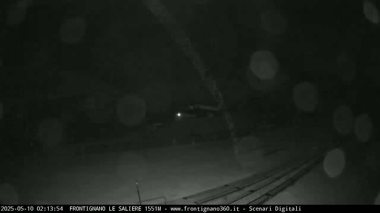 A nighttime, snowy scene shows a dark, empty area with indistinct objects and a set of parallel lines in the foreground.