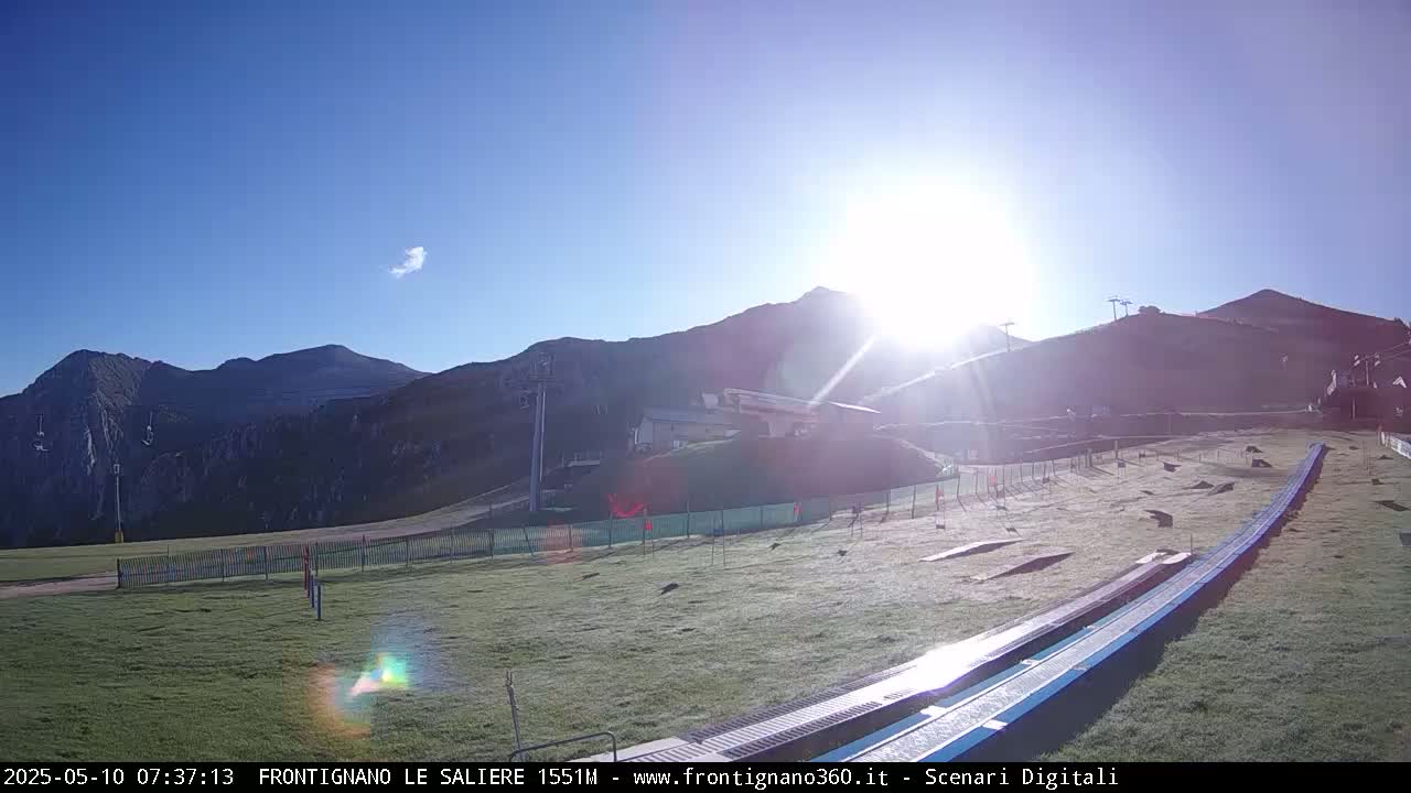 A sunny, clear day reveals a grassy area with a long, blue track leading towards a mountain range, with ski lifts visible on the slopes.
