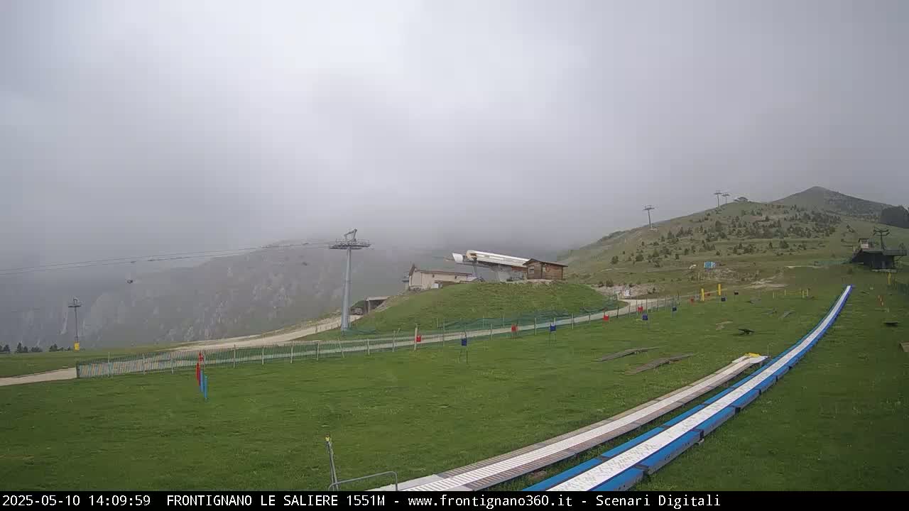 A foggy mountaintop view shows a grassy area with ski lifts, a small building, and two long, flat ramps.