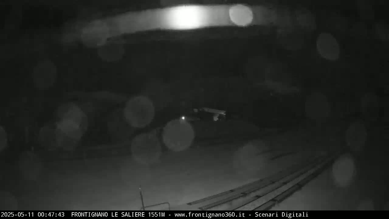 A nighttime, snowy scene shows indistinct objects in the mid-ground and a dark, snowy ground with parallel lines in the foreground.