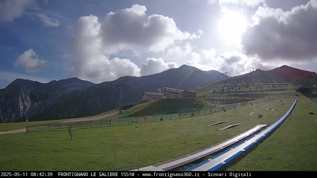 A grassy area with a ski lift and a long, metal ramp leads towards mountainous terrain under a partly sunny sky with fluffy white clouds.