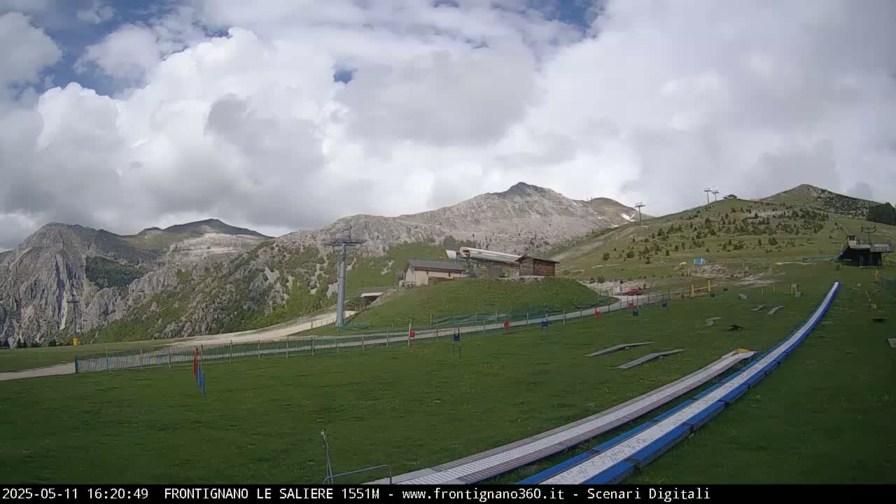 A grassy mountainside with a ski lift, small buildings, and two long ramps, under a partly cloudy sky.