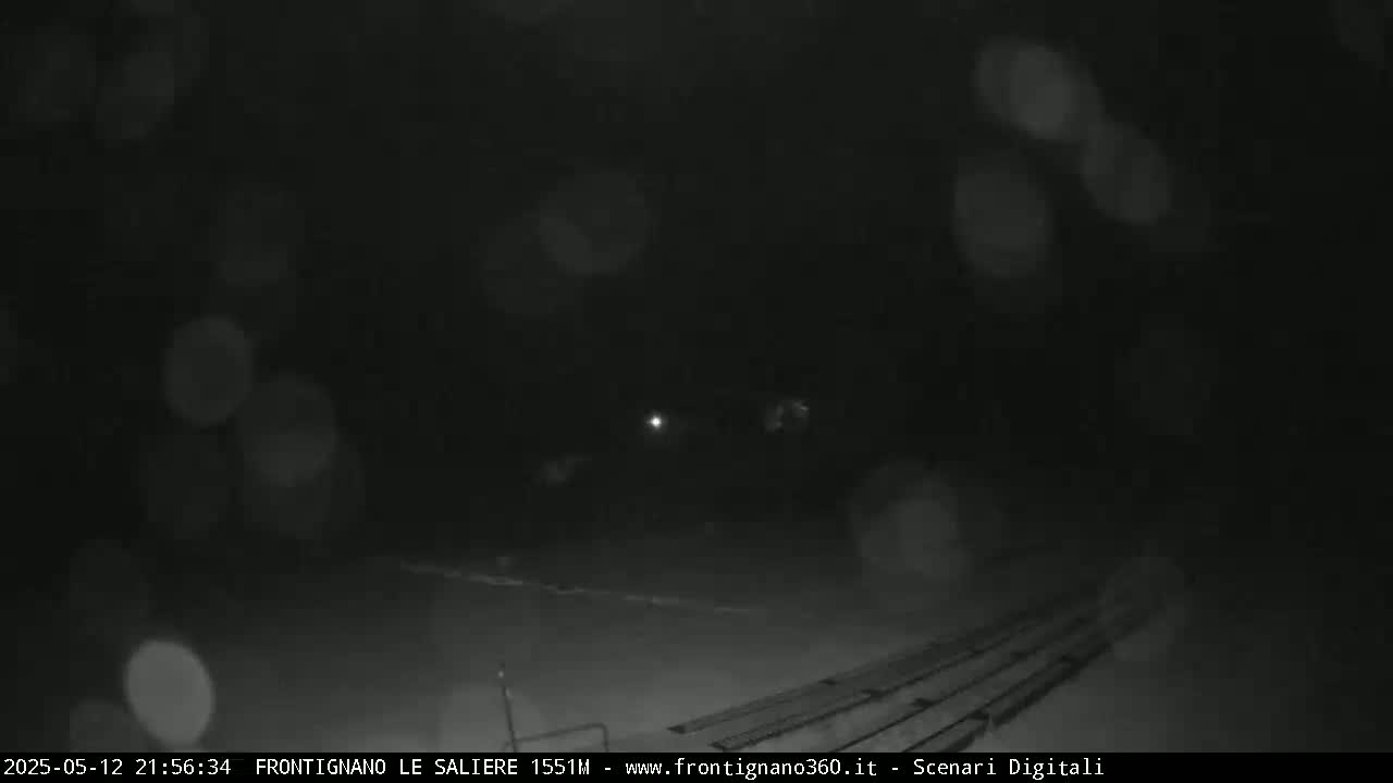A nighttime, snowy scene shows a dark, mostly empty area with faintly visible parallel lines on the ground and blurry lights in the distance.