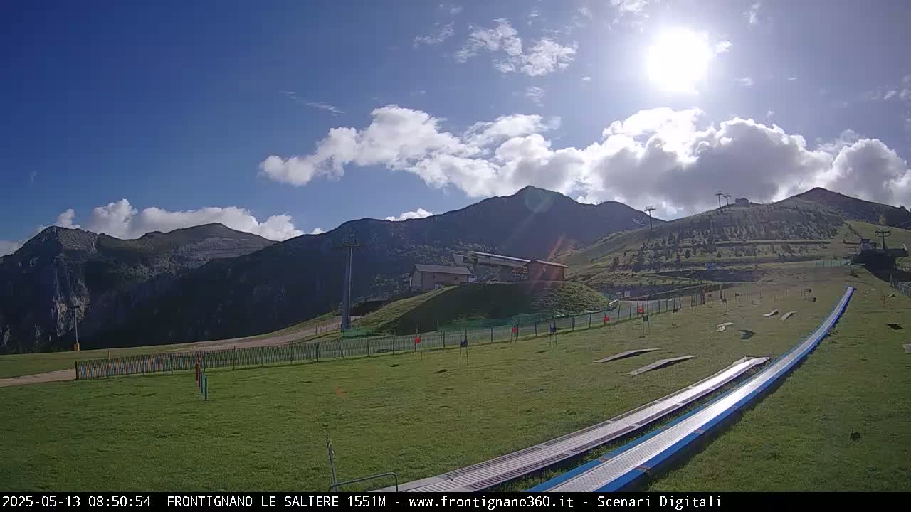 A sunny day shows a grassy mountainside with a ski lift, a small building, and two long, blue ramps leading down the slope.