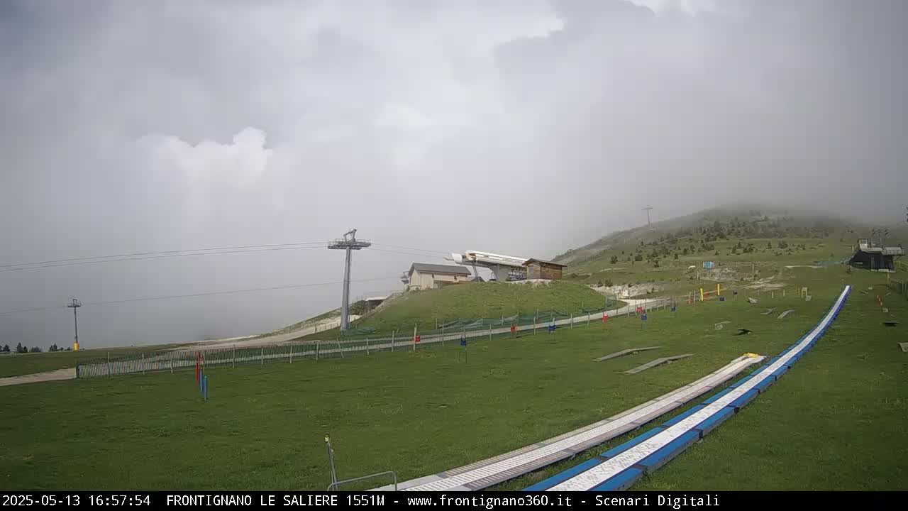 A grassy hill with ski lifts and ramps is shrouded in fog under a cloudy sky.