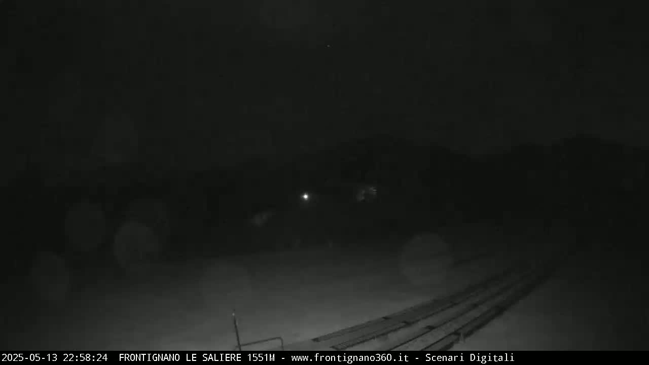A dark, nighttime scene shows a snow-covered ground with faint lights in the distance under a clear sky.