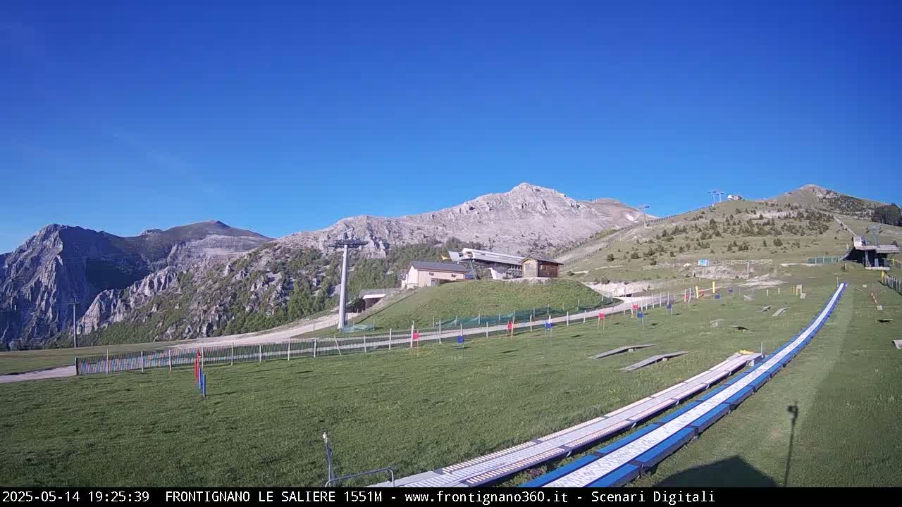 A grassy mountaintop area with a ski lift, small building, and long, flat tracks under a clear blue sky.