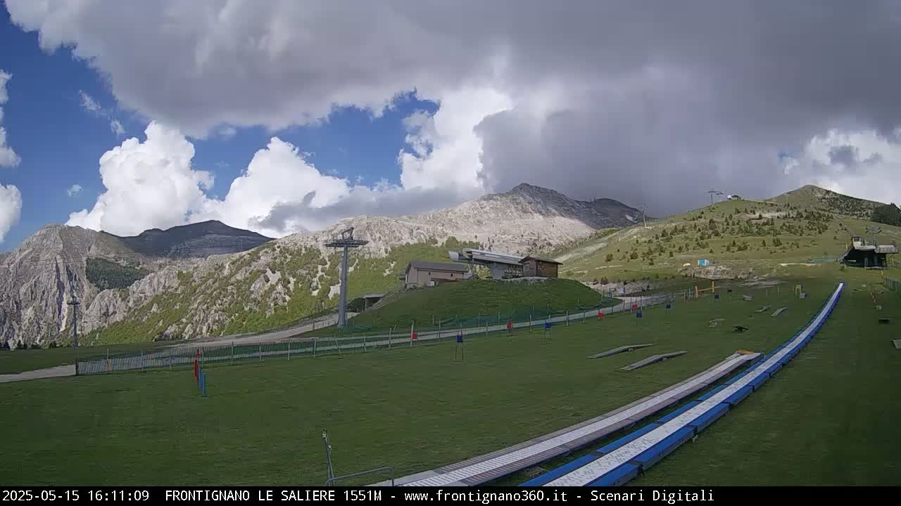 A grassy mountainside with ski lifts, a small building, and a pair of long, flat ramps under a partly cloudy sky.