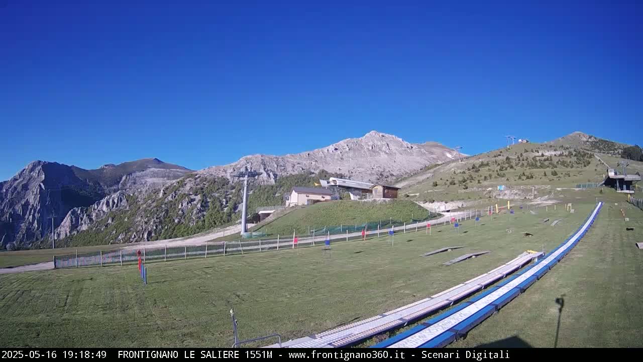 A grassy mountaintop area with a ski lift, small buildings, and a long, flat, blue track under a clear blue sky.