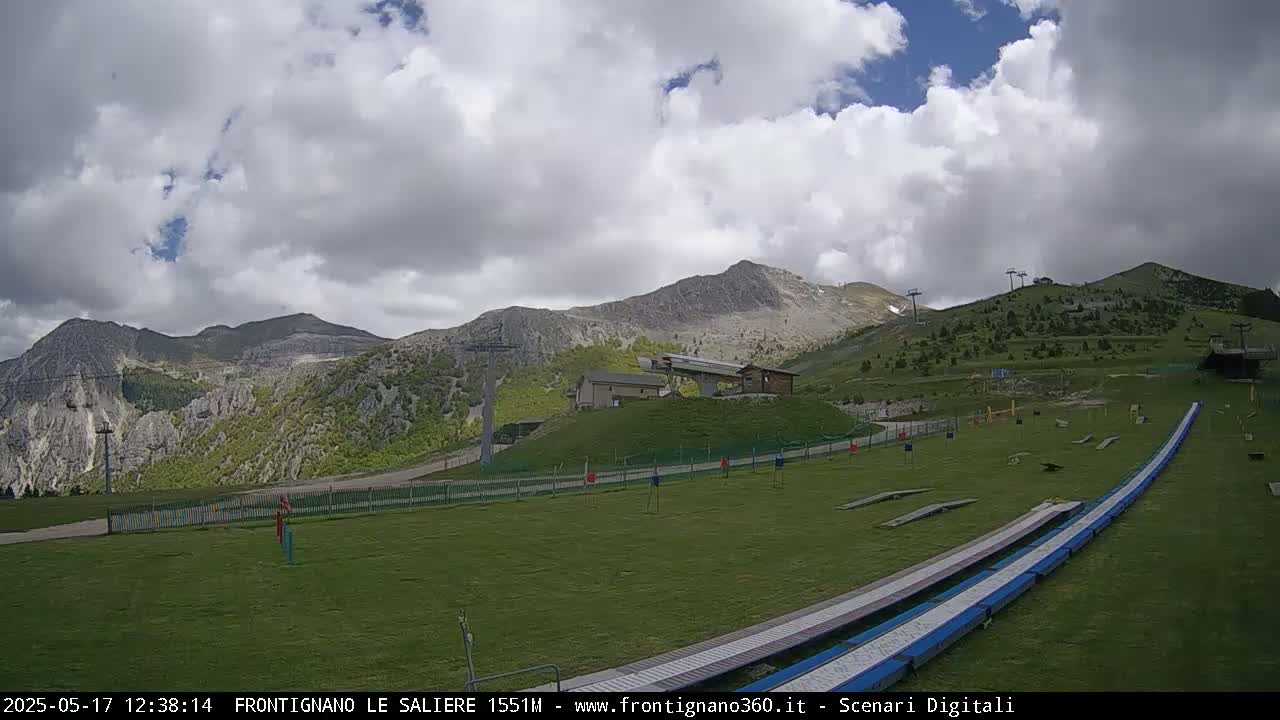 A grassy mountainside with ski lifts and a long, low structure, under a partly cloudy sky.