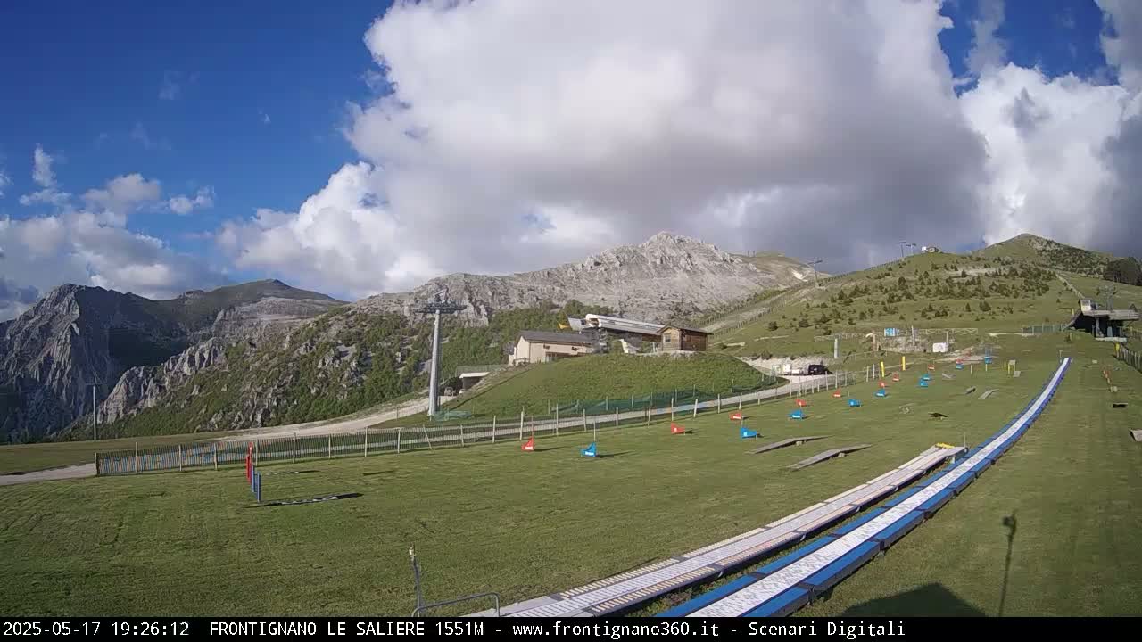 A grassy area with a ski lift and mountainous backdrop under a partly cloudy sky.