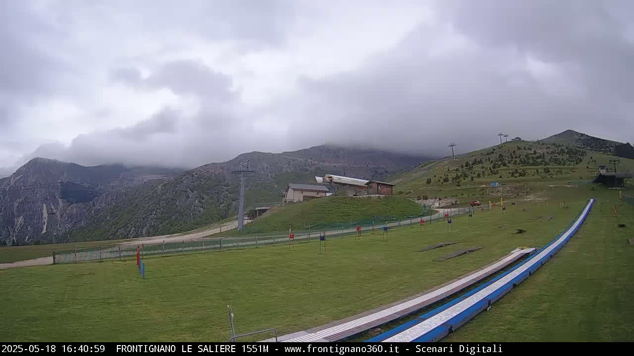 A grassy mountaintop area with a ski lift, a small building, and a long, low slide, under an overcast sky.