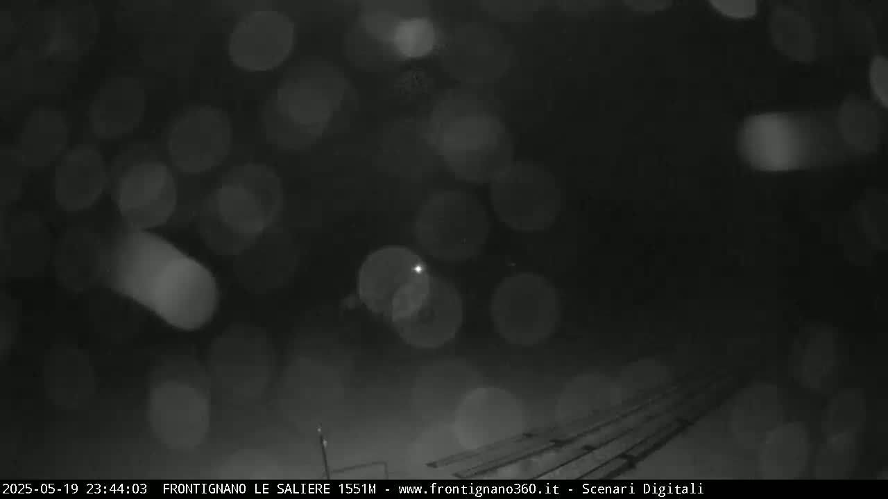 A nighttime, snowy scene shows blurred lights and a dark surface with parallel lines underneath a heavy snowfall.