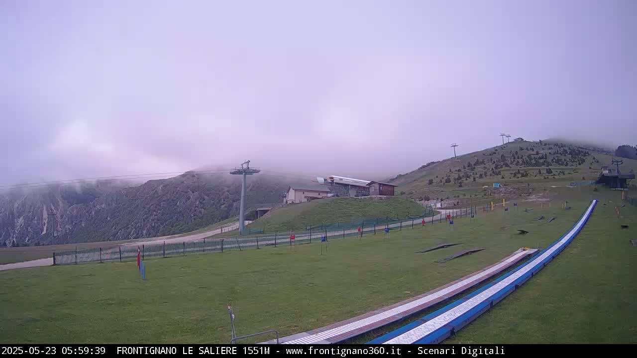A foggy mountain landscape features a grassy area with ski lifts, a small building, and two long, flat ramps.