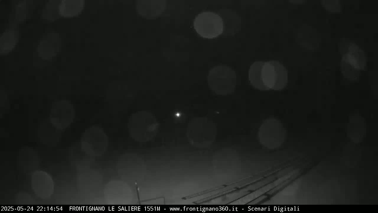 A dark, nighttime scene shows blurry lights in the distance and a rail track in the foreground, with light snowfall or mist.
