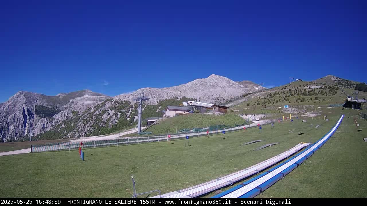 A grassy mountainside with a ski lift, small buildings, and two long, white, and blue ramps under a clear blue sky.
