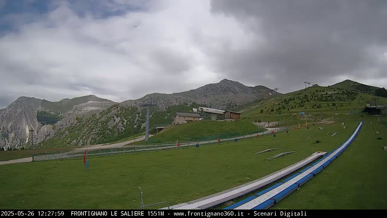A grassy mountainside with ski lifts, a few buildings, and a paved area with what appears to be a summer toboggan run under an overcast sky.