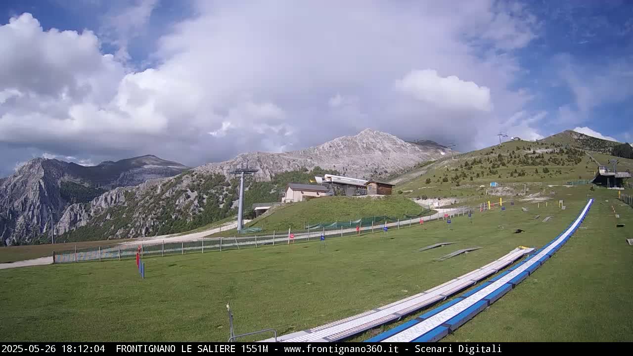 A grassy mountaintop area with a ski lift and a long, flat, blue-and-white track, under a partly cloudy sky.