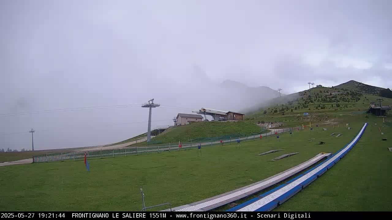 A foggy mountaintop scene features a grassy area with ski jump ramps, chairlift towers, and a small building, all under an overcast sky.
