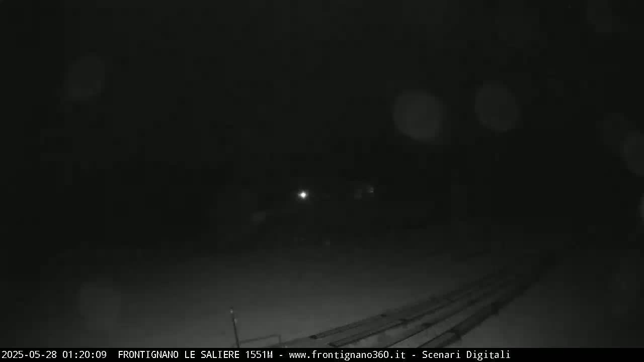 A nighttime, snowy scene shows a dark, mostly empty area with faint lights and parallel lines in the lower portion of the frame.