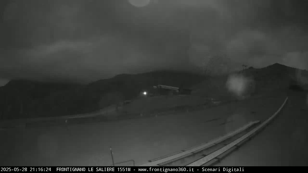 A nighttime, low-light view shows a dark road with parallel lines in the foreground, leading toward dimly lit structures and dark hills under a cloudy sky.