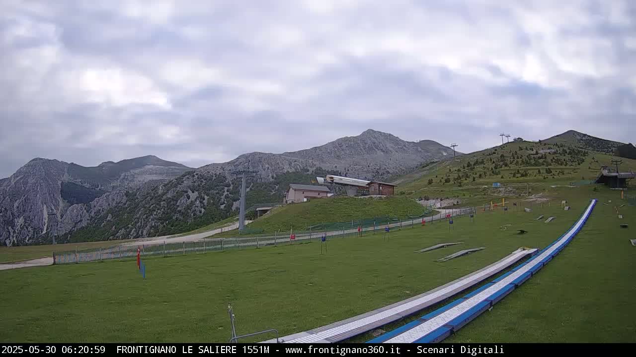 A grassy mountainside area with a ski lift and two long, flat structures, under a cloudy sky.