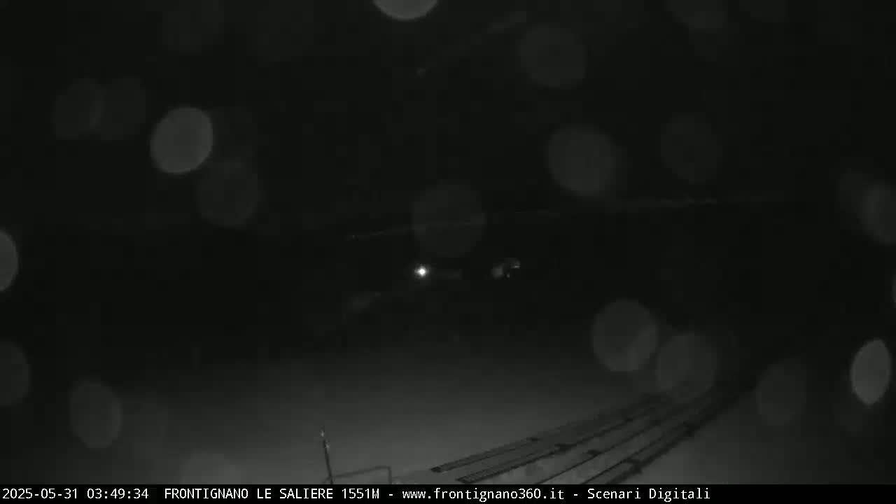 A nighttime, snowy scene shows indistinct shapes and blurry lights in the distance.