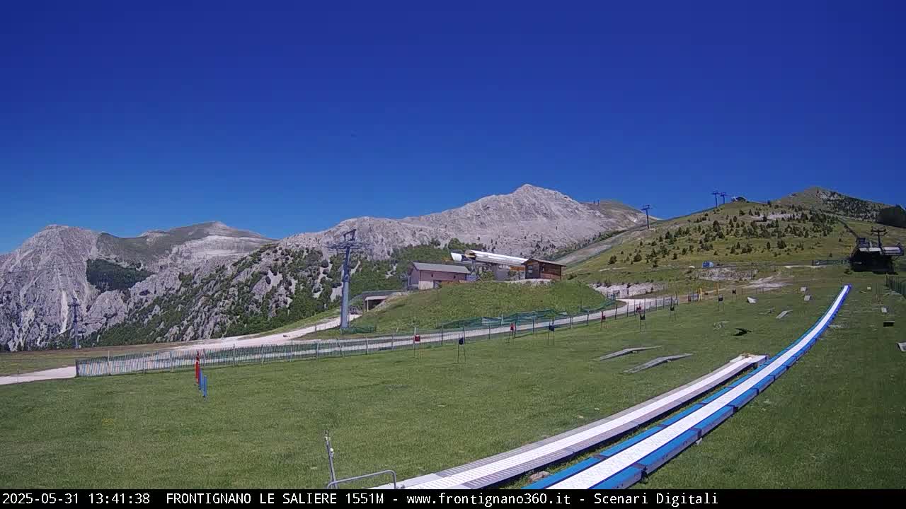 A grassy mountain slope with a ski lift, small buildings, and two long, white structures that appear to be a type of slide or track, under a clear blue sky.