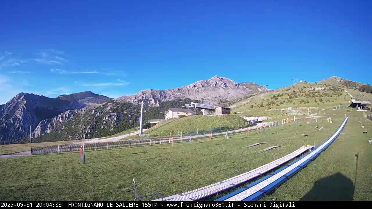 A grassy mountaintop area with a ski lift and long, white ramps, under a clear blue sky.