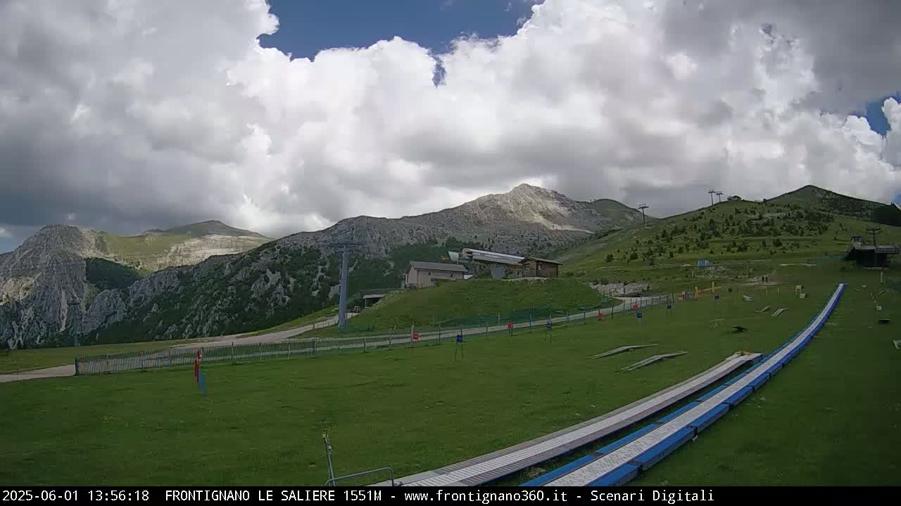 A grassy mountaintop area with ski lifts and a long, flat structure, under a partly cloudy sky.