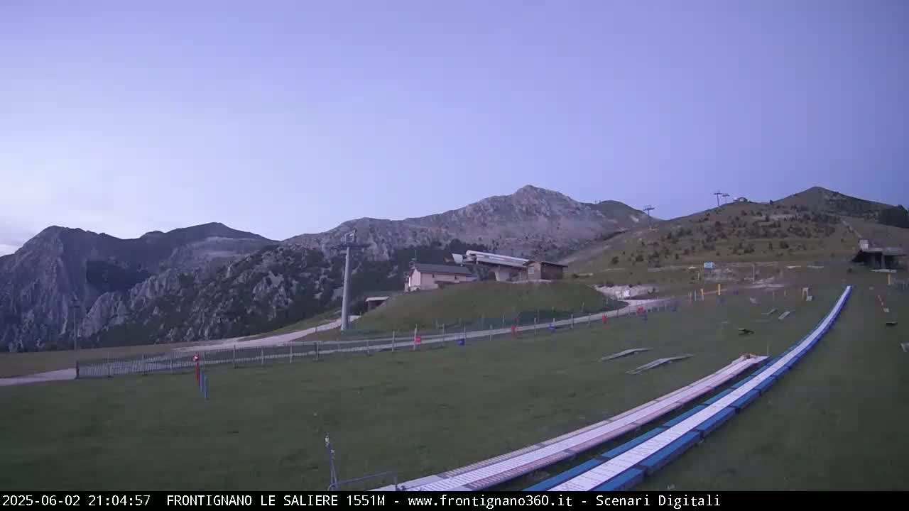 A grassy mountain slope with ski lifts and a pair of long, flat ramps, under a twilight sky.