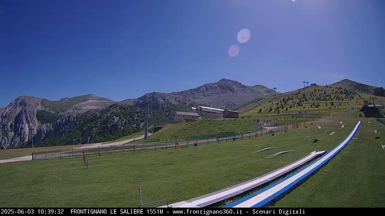 A sunny day reveals a mountain landscape with grassy slopes, ski lifts, and two long, parallel ramps, likely for a summer activity.