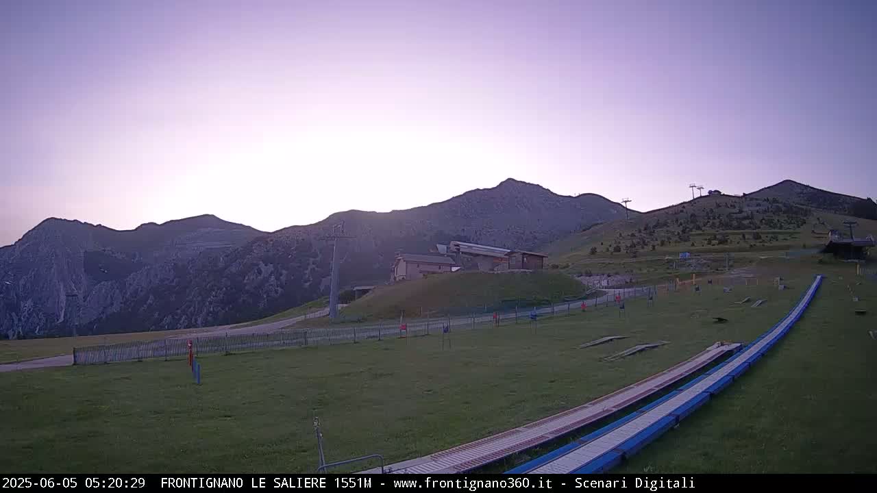 A grassy mountaintop area with a few buildings, ski lifts, and long ramps, bathed in the soft light of either sunrise or sunset under a clear, pale purple sky.