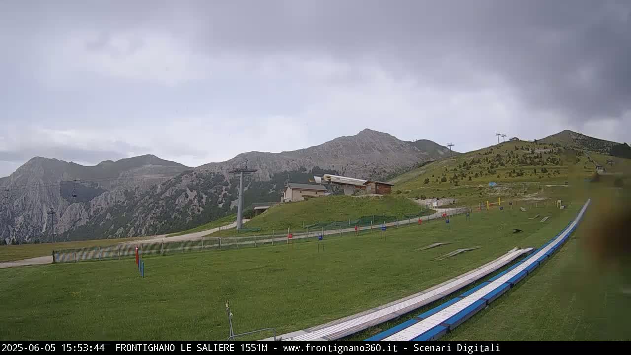 A grassy mountain slope with ski lifts, a small building, and a long, light-blue track stretches towards the foreground under an overcast sky.