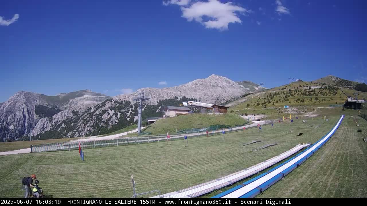 A grassy mountaintop area with a long, white slide, a small building, and a mountain range in the background under a clear blue sky.
