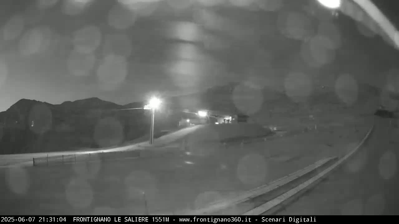 A nighttime, rainy view of a road curving toward a mountain range under artificial light.