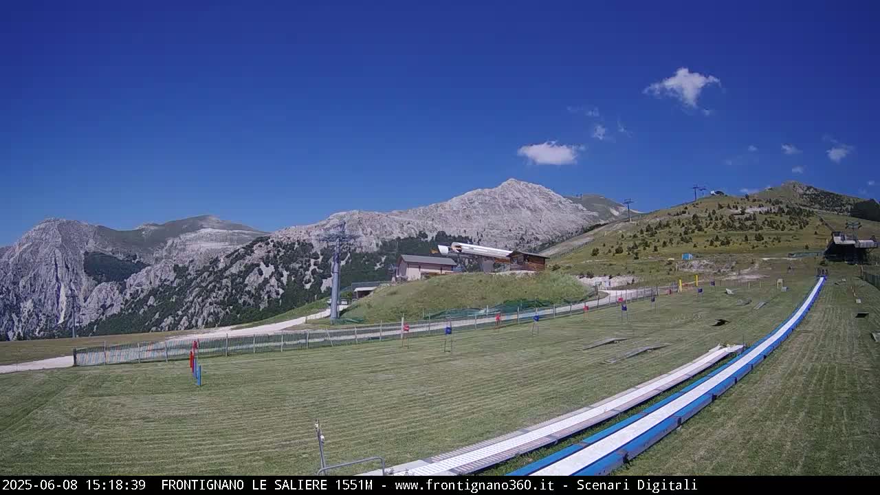 Under a clear blue sky, two long, white slides are situated on a grassy field, with a mountain range and ski lift infrastructure in the background.